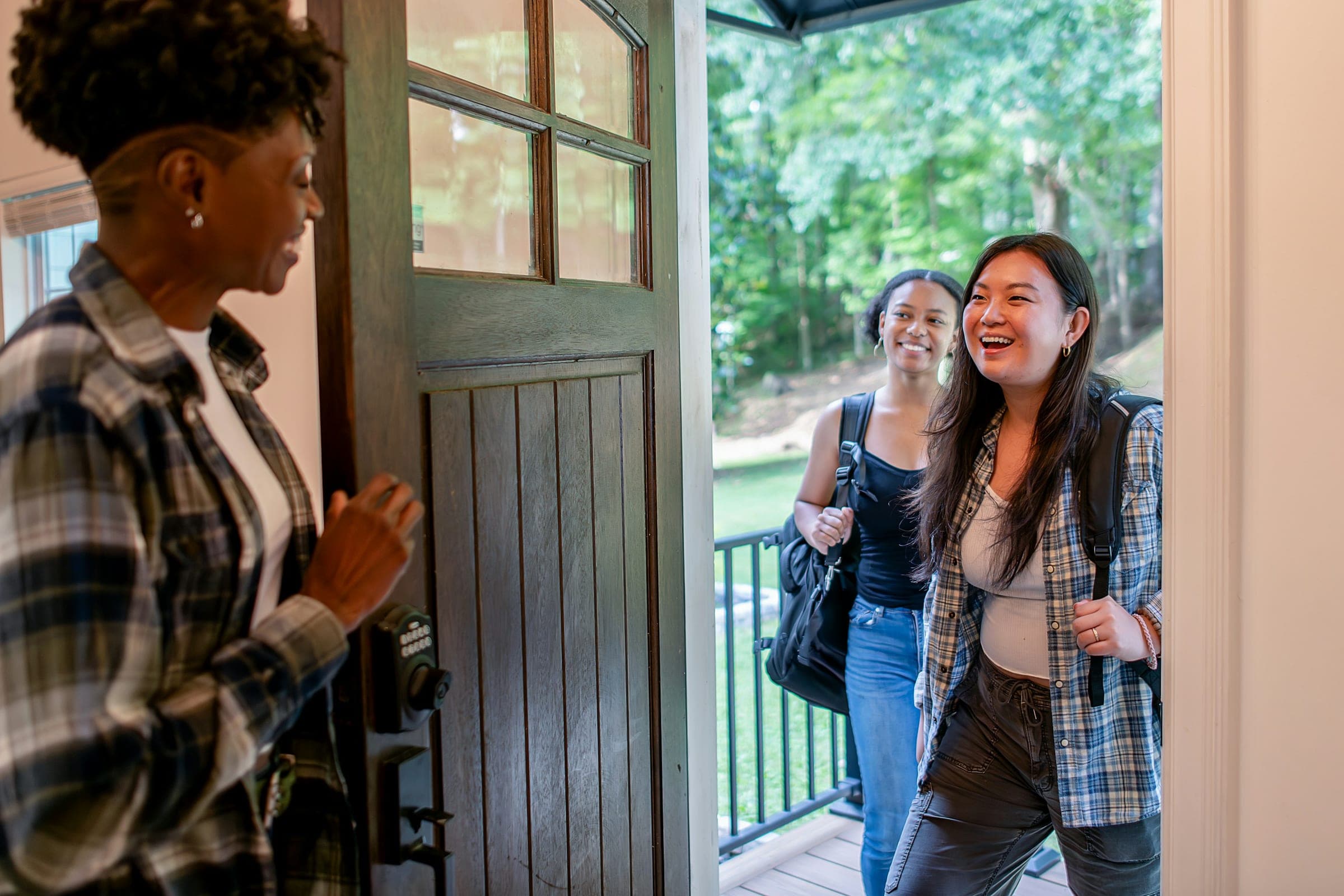 A smiling host opening the front door to two happy guests with backpacks on a sunny day