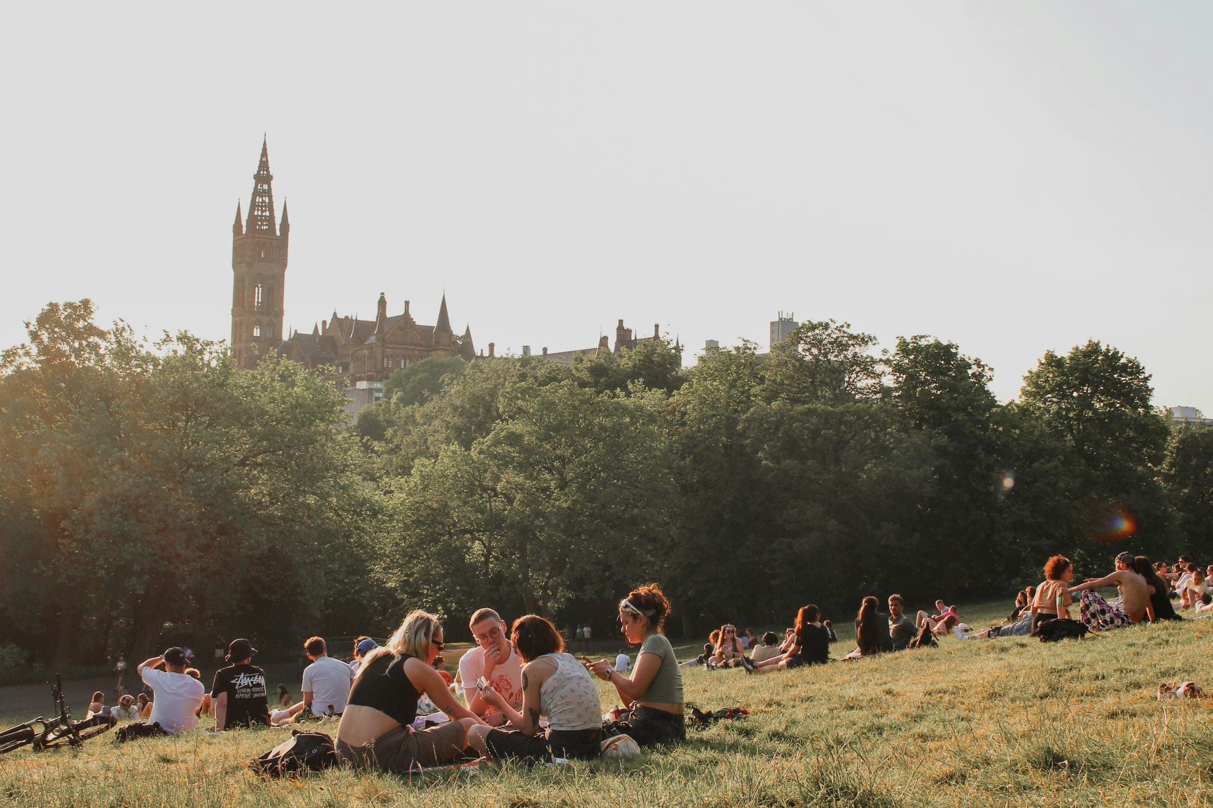 Friends sitting on the grass in Kelvingrove Park on a sunny evening, with the University of Glasgow’s tower beyond the trees