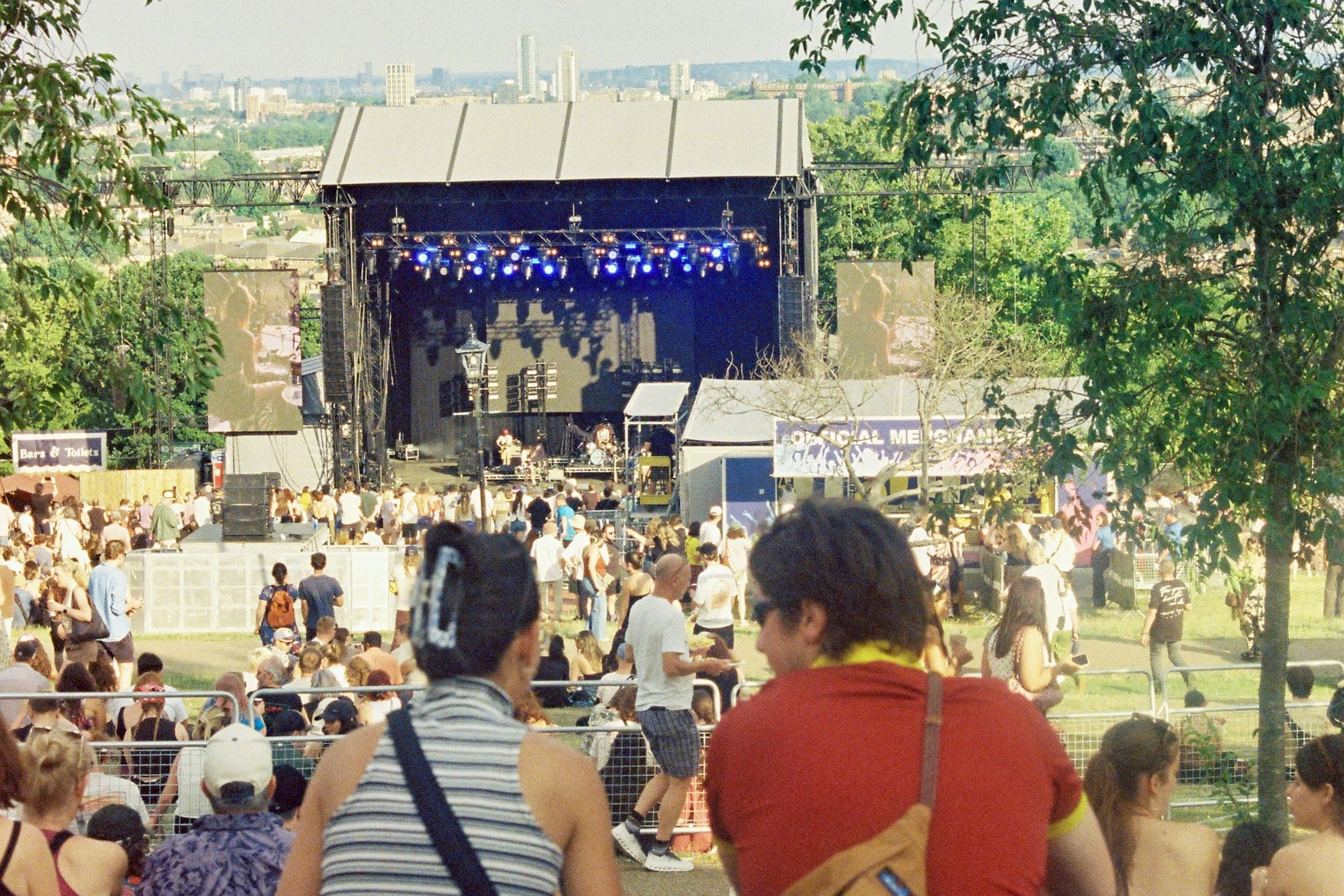 Two friends sitting on a grassy slope watching an open-air stage in sunny parkland, with trees around and a crowd below